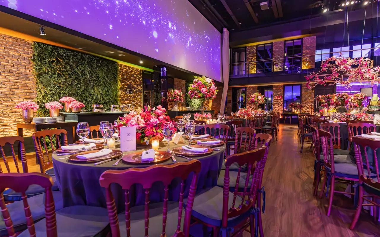 A banquet hall set for a seated dinner, round tables with tall floral centerpieces under colored uplighting