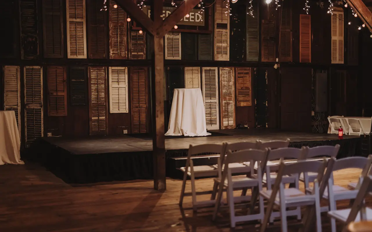 The inside of a barn venue set for a ceremony, white folding chairs facing a low stage under string lights