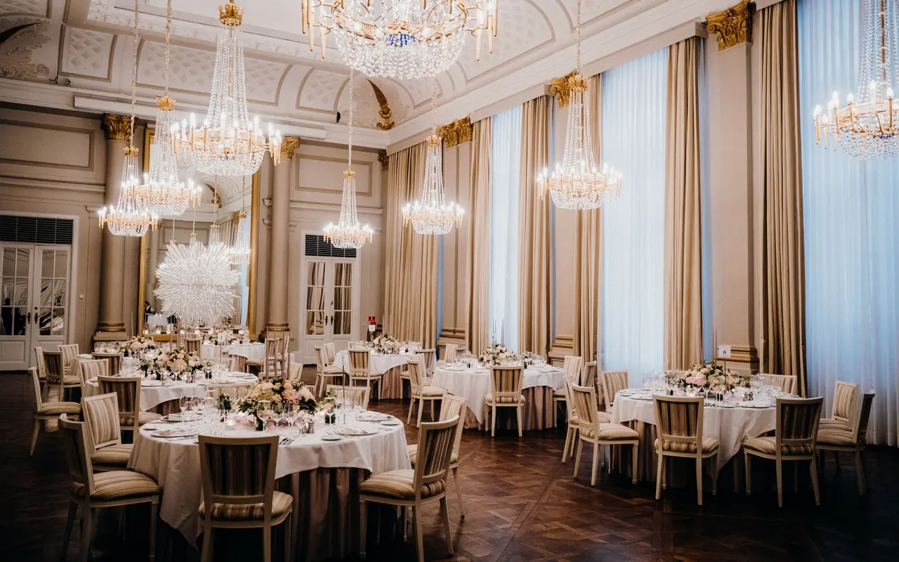 A hotel ballroom with chandeliers and tall draped windows, set with round tables for a seated event