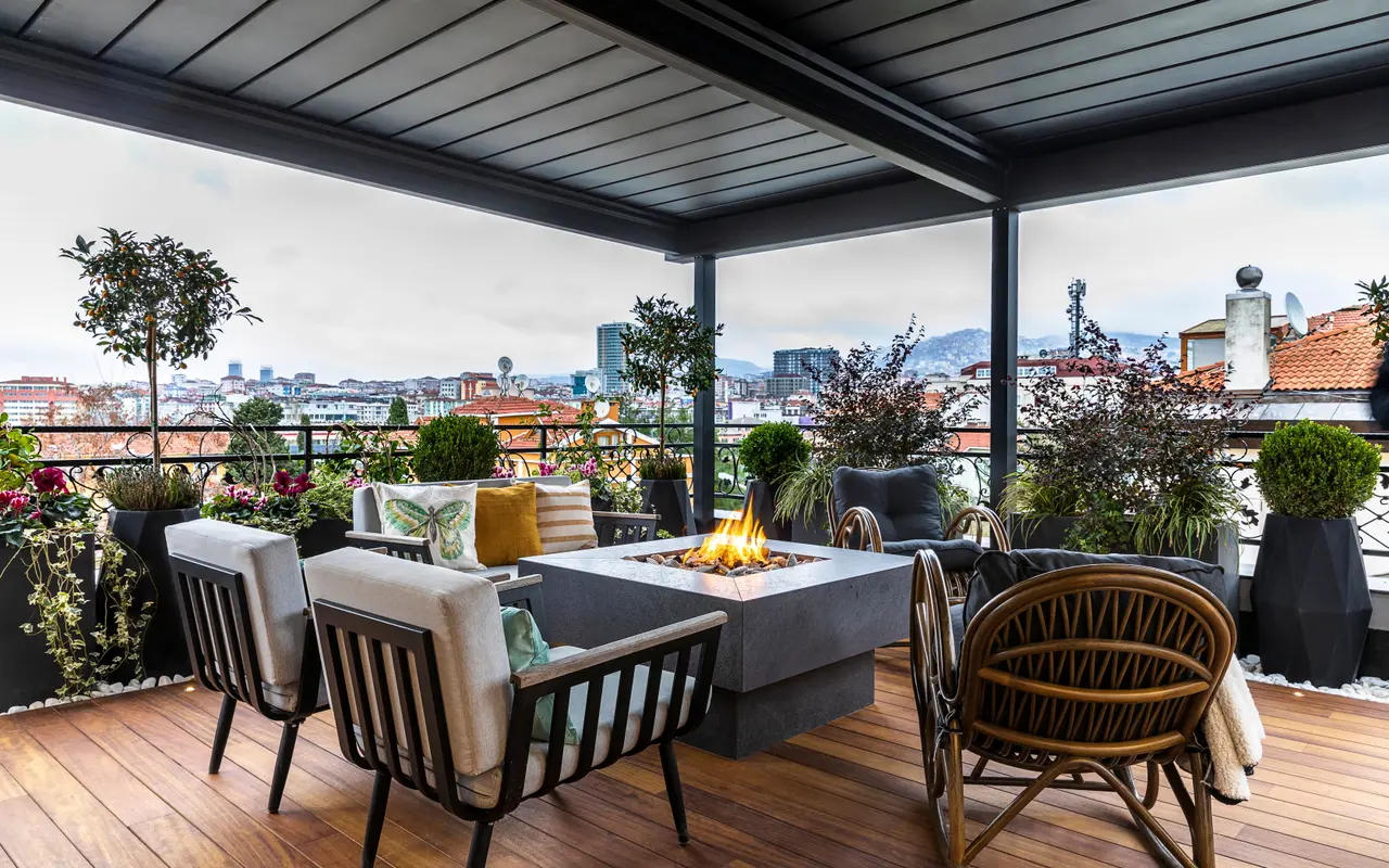 A covered rooftop terrace with lounge seating and a fire table, city rooftops behind it